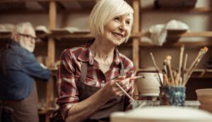Woman painting clay pot with senior potter at workshop